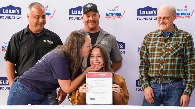 A smiling young woman being kissed by her mom after signing on to a skilled career. Three men stand behind smiling.