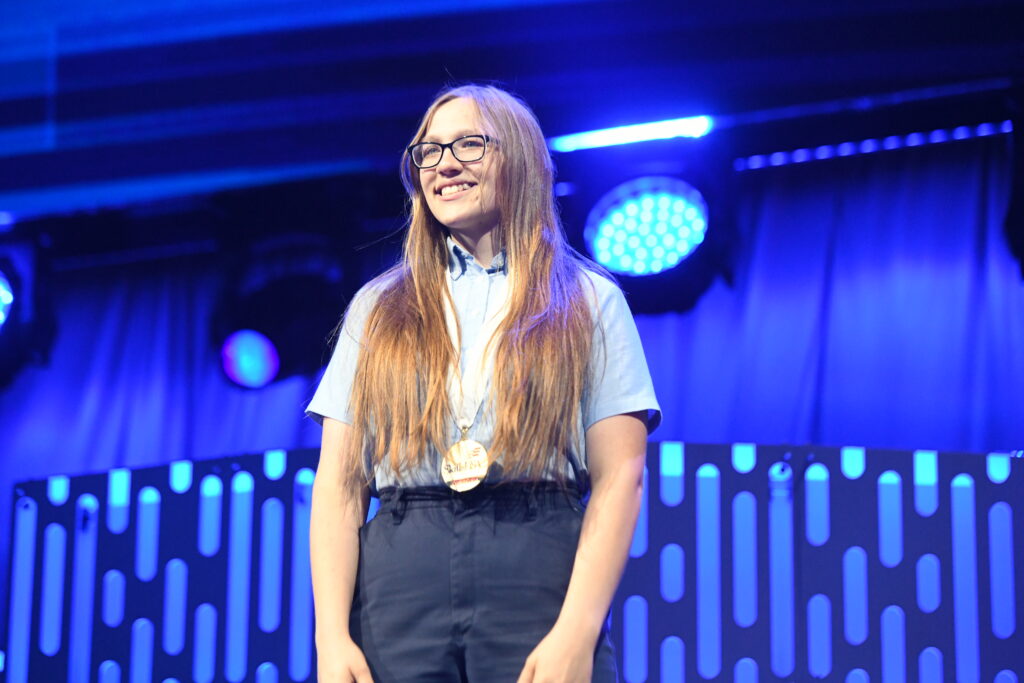 A person with long hair and glasses stands on a stage under blue lighting, smiling while wearing a medal around their neck.