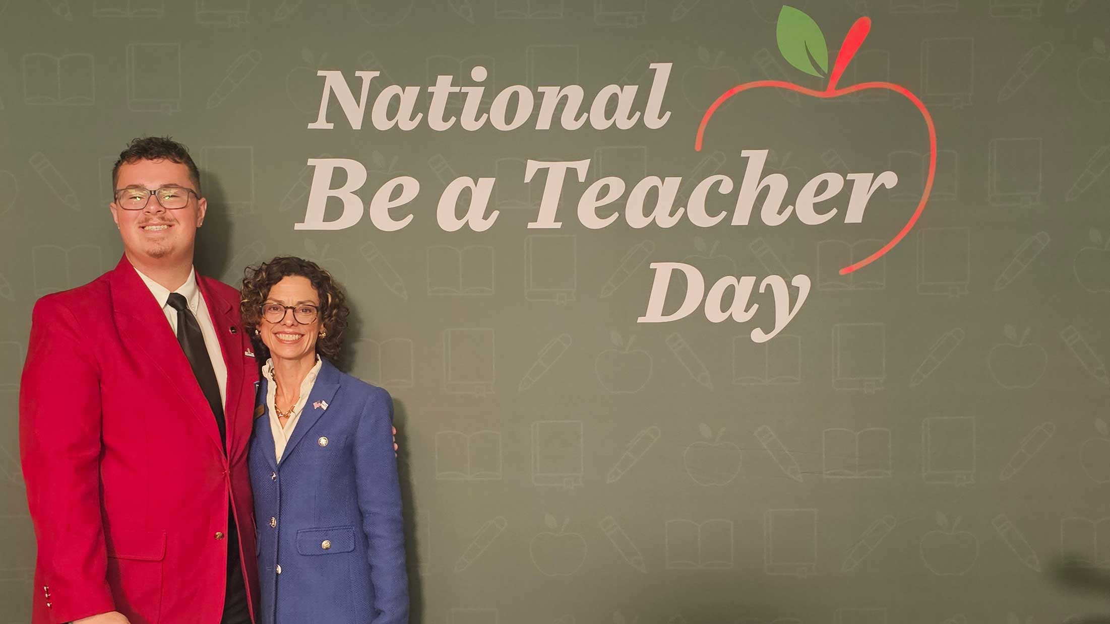 Curtis Muecke in an official red blazer poses with Chelle Travis. She's wearing a blue blazer in front of a green National Be a Teacher Day backdrop decorated with an apple logo.