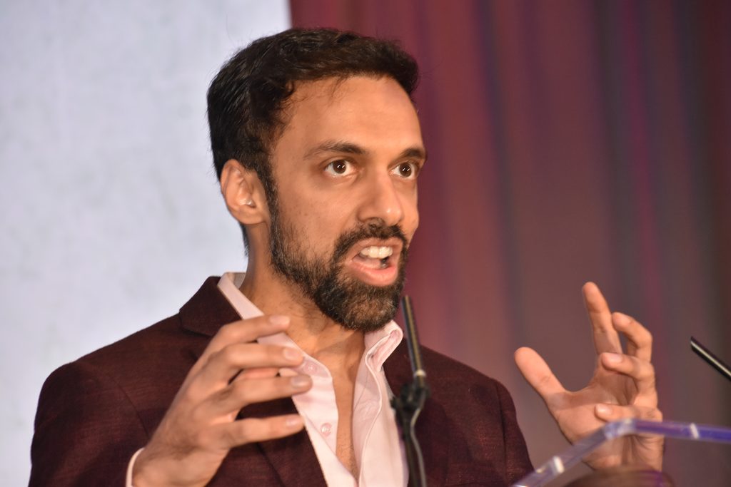 A person with dark hair and a beard, wearing a maroon blazer over a light-colored shirt, is speaking at a podium while gesturing with both hands.